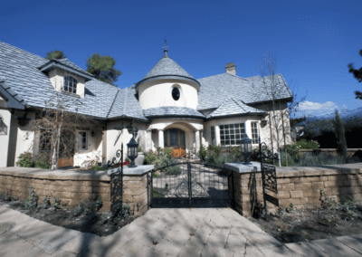 Elegant two-story residence with a turreted entryway and slate roof, complemented by a stone retaining wall and wrought-iron gate.