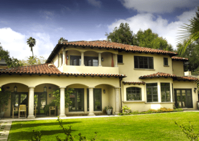 Spanish-style two-story house with arches and a tile roof, surrounded by lush greenery.