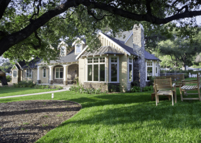 Suburban house with a large windowed sunroom and a manicured lawn, shaded by mature trees.