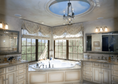 A luxurious bathroom with a large tub, dual vanities, elegant draperies, and a chandelier hanging from a decorative ceiling.