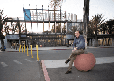 A person sitting on a large planter in front of the Los Angeles Zoo entrance with palm trees and a clear sky in the background.