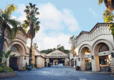 Entrance of an international marketplace with archways, palms, and a sign overhead. Pathway leads to various shops and greenery on both sides. Sunny sky above.