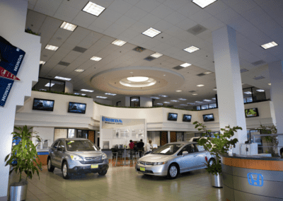 A modern car dealership showroom featuring two cars on display, a sales desk, and several wall-mounted televisions.