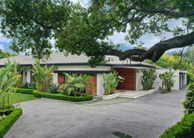 Modern single-story house with wood and white exterior, surrounded by lush greenery and trees, featuring a spacious driveway.