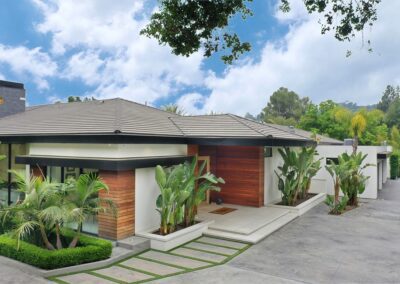 A modern house with a flat roof, large windows, and wood accents, surrounded by tropical plants and a paved driveway, set against a backdrop of trees and a partly cloudy sky.