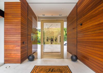 Modern entrance with wooden panel walls, a large glass door, and a doormat with a letter "H". Two black spherical decorations sit at the base of the door.