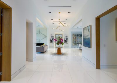 Spacious modern hallway with glossy white tile floor, abstract light fixture, and a central table with a floral arrangement. Artwork and open doorways lead to adjoining rooms.