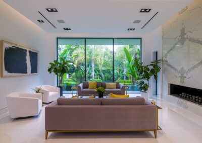 Modern living room with large windows, gray sofas, white armchairs, green plants, and a marble fireplace. A large abstract painting hangs on the wall.