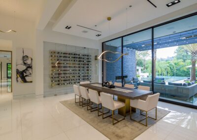 Modern dining room with a long table, white chairs, wine display wall, and a large window view of the patio and garden. Contemporary light fixture hangs above the table.