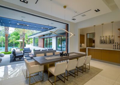 Modern dining area with a wooden table, white chairs, and a contemporary light fixture. Large glass doors open to a patio with outdoor seating and a view of a garden.