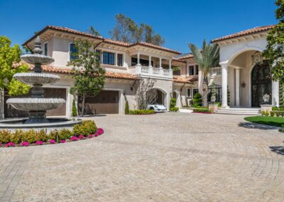 A large Mediterranean-style mansion with stone driveway, multiple arched entrances, balconies, and a three-tiered fountain surrounded by flowers in the foreground.