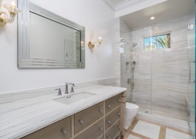 Modern bathroom with a marble countertop, large mirror, glass shower enclosure, and beige cabinets. Sconces are mounted on the wall.