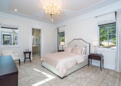 A bright bedroom with a double bed, two bedside tables with lamps, a dresser, a chair, and a chandelier. Two windows with curtains let in natural light. Carpeted floor and white walls.