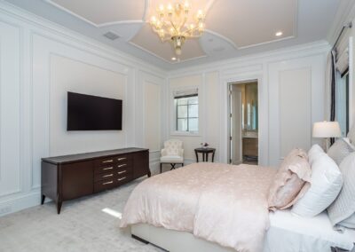 Elegant bedroom with a plush bed, wall-mounted TV, chandelier, and a side table. A door leads to an adjoining room, and a window allows natural light in. White paneled walls add a classic touch.