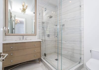 Modern bathroom with a glass-enclosed shower, marble walls, a wooden vanity with a mirror above, and a toilet.