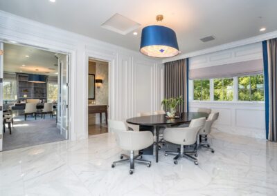 A modern conference room with a round table, six chairs, marble floors, large windows, and a blue chandelier. Another room with a table and chairs is visible through an open door.