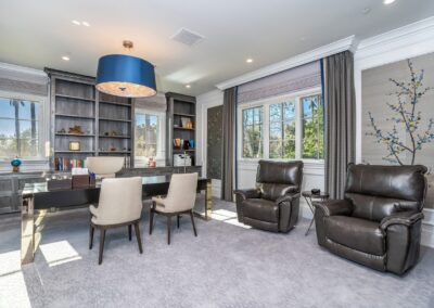 A modern home office with a large desk, two chairs, leather recliners, a bookshelf, and a blue ceiling light. Two windows with curtains allow natural light in.