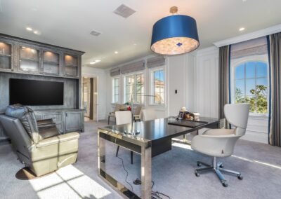 A modern office room with a desk, chairs, a large TV, and leather recliner. A chandelier hangs from the ceiling, and two large windows offer a view outside.