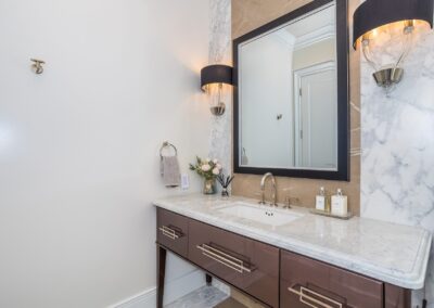 A bathroom with a marble countertop, dark wood vanity, large mirror, two wall sconces, and decorative items on the counter. Towels hang to the left.