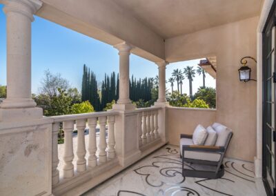 Balcony with white balustrades and a cushioned chair overlooks tall trees and a garden under a clear blue sky.