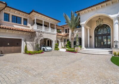 A large luxury house with arched entry, palm trees, and a driveway. A white car is parked near the garage doors.