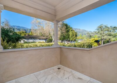 View from a balcony featuring marble flooring, overlooking a garden with trees and a house. Hills and a clear blue sky are visible in the background.