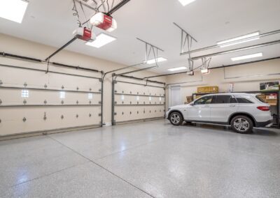 Spacious garage with polished floor, containing a parked SUV and storage shelves on the back wall. Ceiling lights and automatic garage door openers are visible.