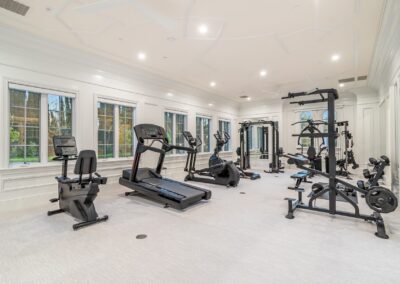 A spacious home gym with various equipment, including a treadmill, elliptical, weight machines, and windows lining one wall.