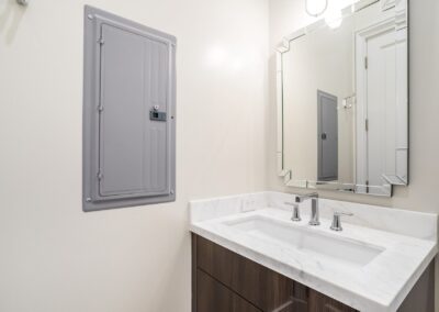 Modern bathroom with a rectangular mirror above a marble countertop sink and a gray electrical panel on the adjacent wall.