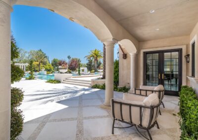 Outdoor patio with cushioned chairs under arches, overlooking a sunlit pool and landscaped garden with palm trees.