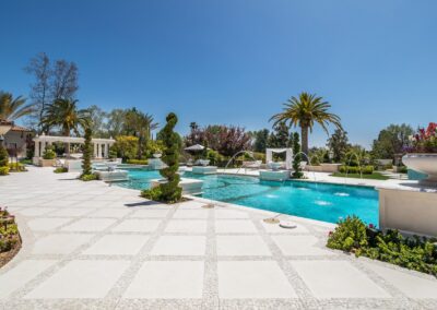 Luxurious outdoor pool area with fountains, surrounded by manicured gardens, palm trees, and a spacious tiled patio under a clear blue sky.