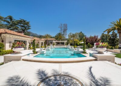 Luxurious mansion backyard with large pool, palm trees, and decorative planters, under a clear blue sky.