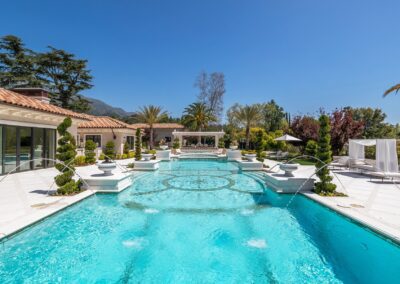 Luxurious outdoor pool surrounded by decorative fountains, lounge chairs, and landscaped greenery under a clear blue sky.