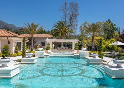 Luxurious backyard with a large swimming pool, surrounded by palm trees and elegant landscaping, with a spacious patio and lounge area in the background under a clear blue sky.