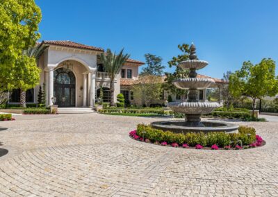 Large mansion with Mediterranean architecture, featuring a circular driveway and a multi-tiered stone fountain, surrounded by vibrant landscaping and clear blue sky.