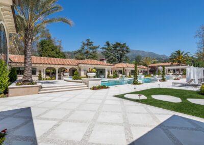 Luxurious outdoor area with a large pool, palm trees, manicured gardens, and terracotta-roofed buildings under a clear blue sky.