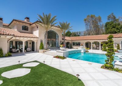 Luxury house with terracotta roof, a pool, and palm trees in the backyard. The architecture features large arched windows and light stucco walls.