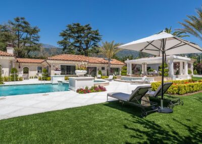 A luxurious backyard featuring a pool, lounge chairs, and a large umbrella. The scene is surrounded by green grass, trees, and a villa with terra cotta roof tiles.