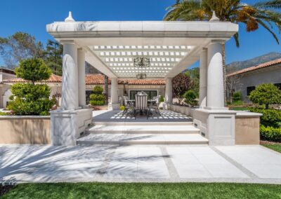 Pergola with dining table and chairs on a patio, surrounded by lush greenery, against a backdrop of a white building and blue sky.