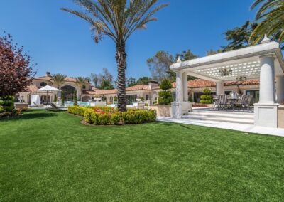 Spacious backyard with a lawn, palm trees, and a covered outdoor seating area. Mediterranean-style house in the background under a clear blue sky.