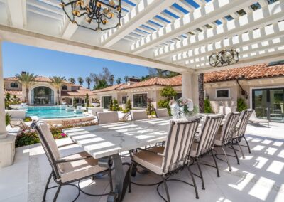 Outdoor patio with a long dining table, chairs, and a pergola. In the background, there's a pool, palm trees, and a large house with terracotta roof tiles.