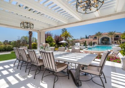 Outdoor patio with a long dining table and chairs under a pergola. A pool and landscaped garden with palm trees are in the background, and a large villa is visible beyond the pool.