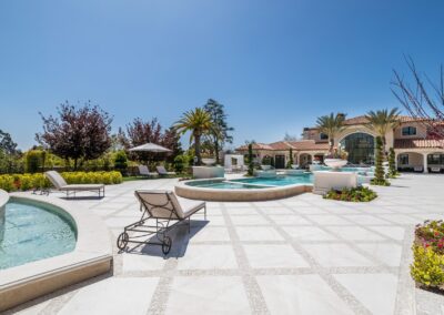 Luxurious mansion with a large pool area, lounge chairs, palm trees, and manicured gardens under a clear blue sky.