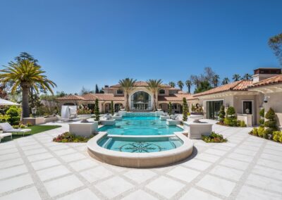 Luxurious mansion with a large, ornate pool surrounded by palm trees and manicured gardens under a clear blue sky.