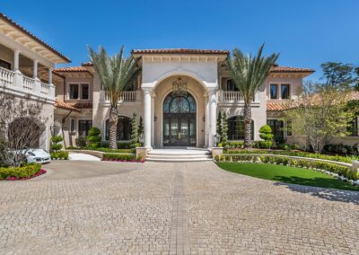 A large, elegant mansion with a stone driveway, archway entrance, and landscaped gardens under a clear blue sky.