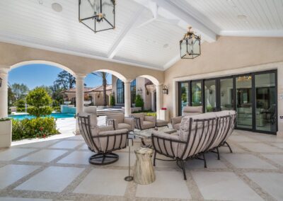 Outdoor patio with cushioned seating set, under a white ceiling. Arched openings lead to a garden with a pool. Glass doors on the right.