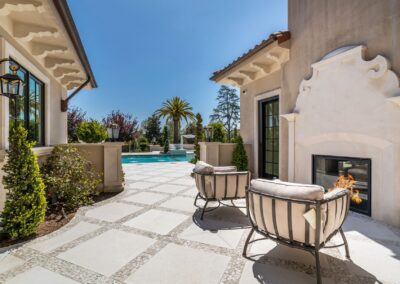 A sunny patio with two cushioned chairs facing a fireplace. Beyond is a pool surrounded by trees and palm, with clear skies above.