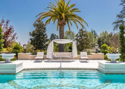 Luxurious outdoor pool area with white lounge chairs and a canopy on a sunny day, surrounded by lush greenery and a tall palm tree in the background.