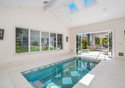 Indoor pool with tiled walls and ceiling skylights. Large windows and open doors show a garden and patio outside.