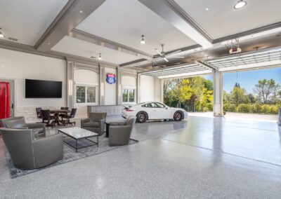 A spacious garage with a white sports car, chairs, a table, a Route 66 sign, and a large TV. The garage door is open, revealing trees outside.